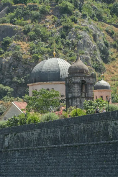 Portretni prikaz drevnih gradskih zidina i crkvenih kupola Kotora, Crna Gora, na pozadini bujnog planinskog pejzaža.