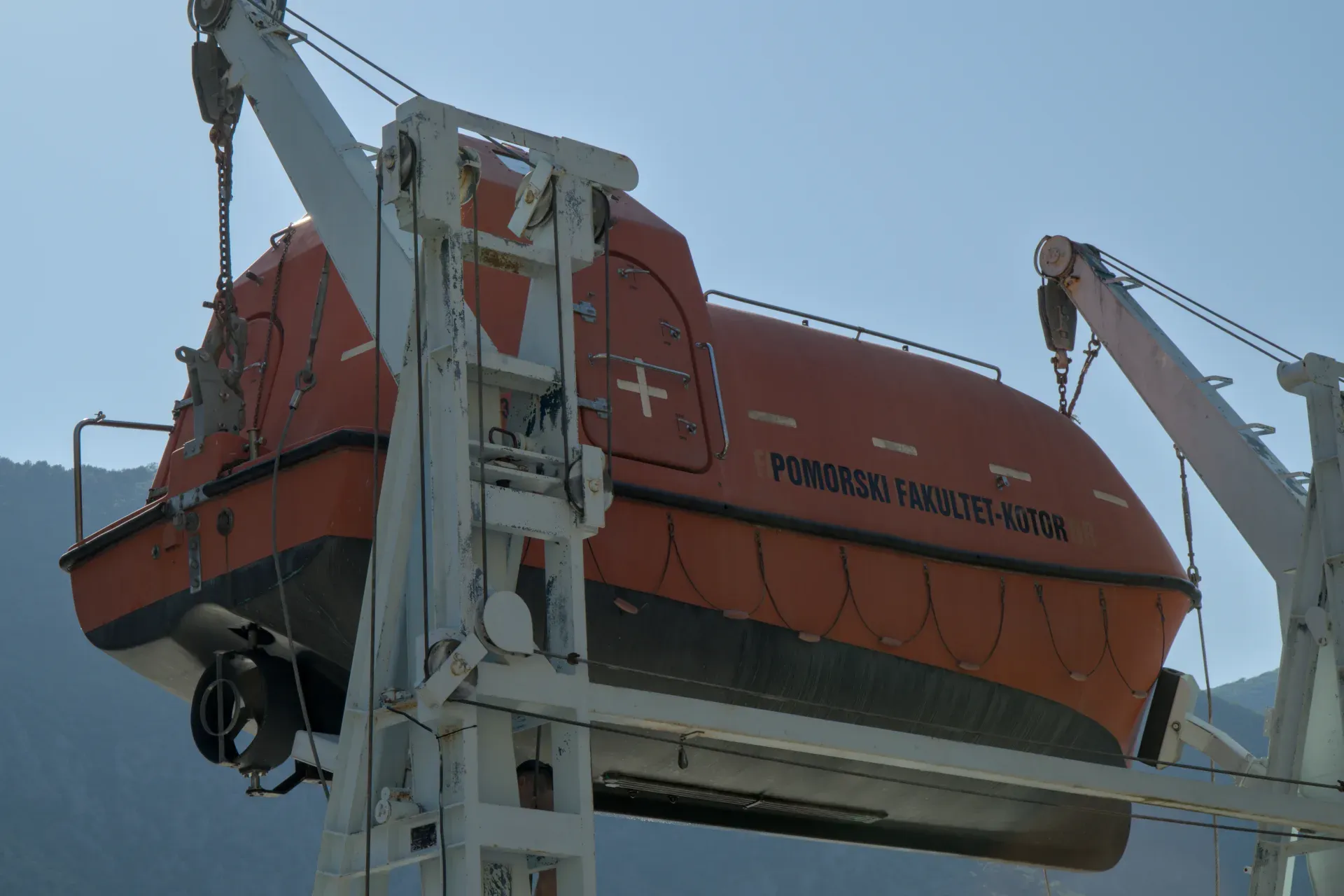 Ein oranges Rettungsboot hängt an einem Kran über dem Wasser, mit blauem Himmel im Hintergrund.