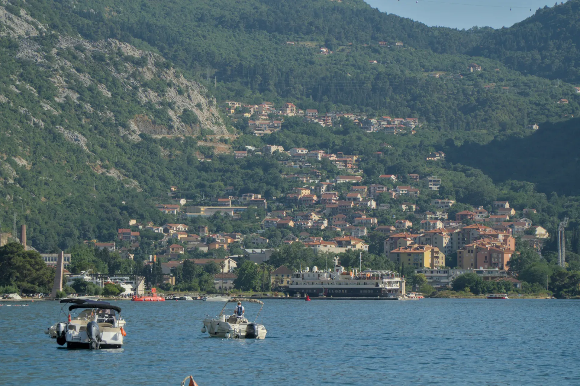 Blick auf die Bucht von Kotor mit Booten auf dem Wasser und einer Stadt, die sich an grüne Berghänge schmiegt.