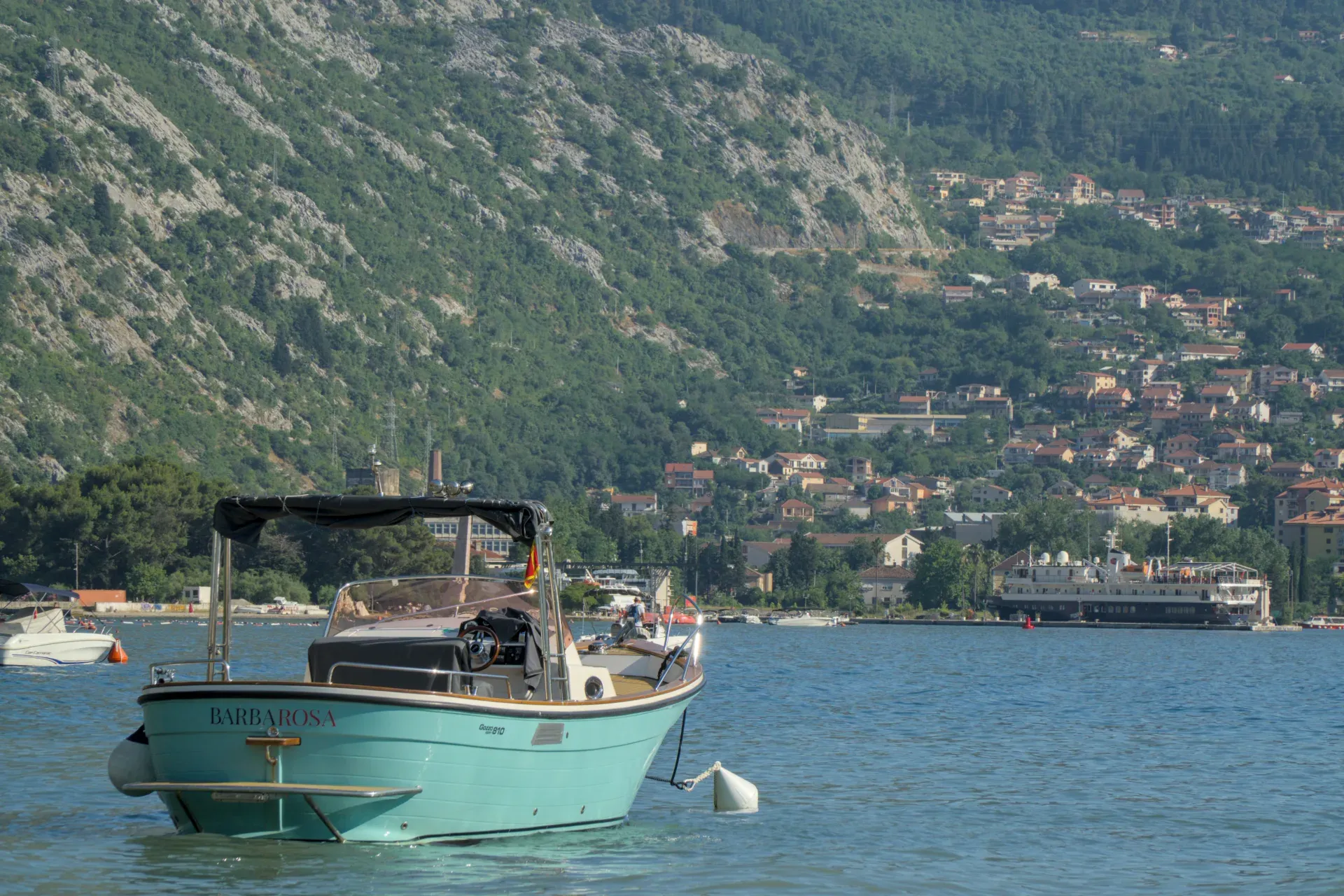 Un barco turquesa descansa frente a la costa de la Bahía de Kotor, Montenegro, con colinas verdes al fondo.