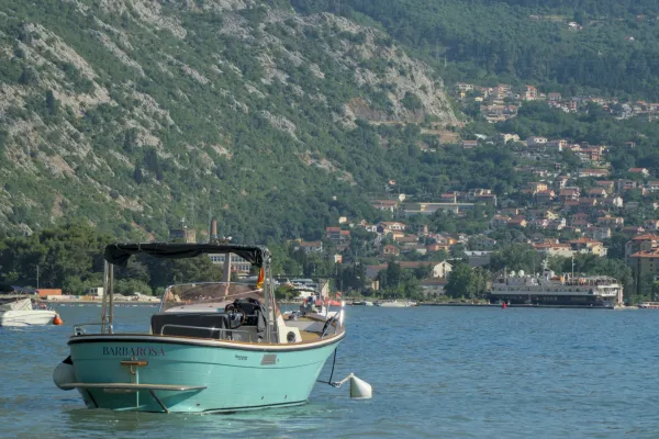 Tirkizni čamac odmara se uz obalu Kotorskog zaliva, Crna Gora, sa zelenim brdima u pozadini.
