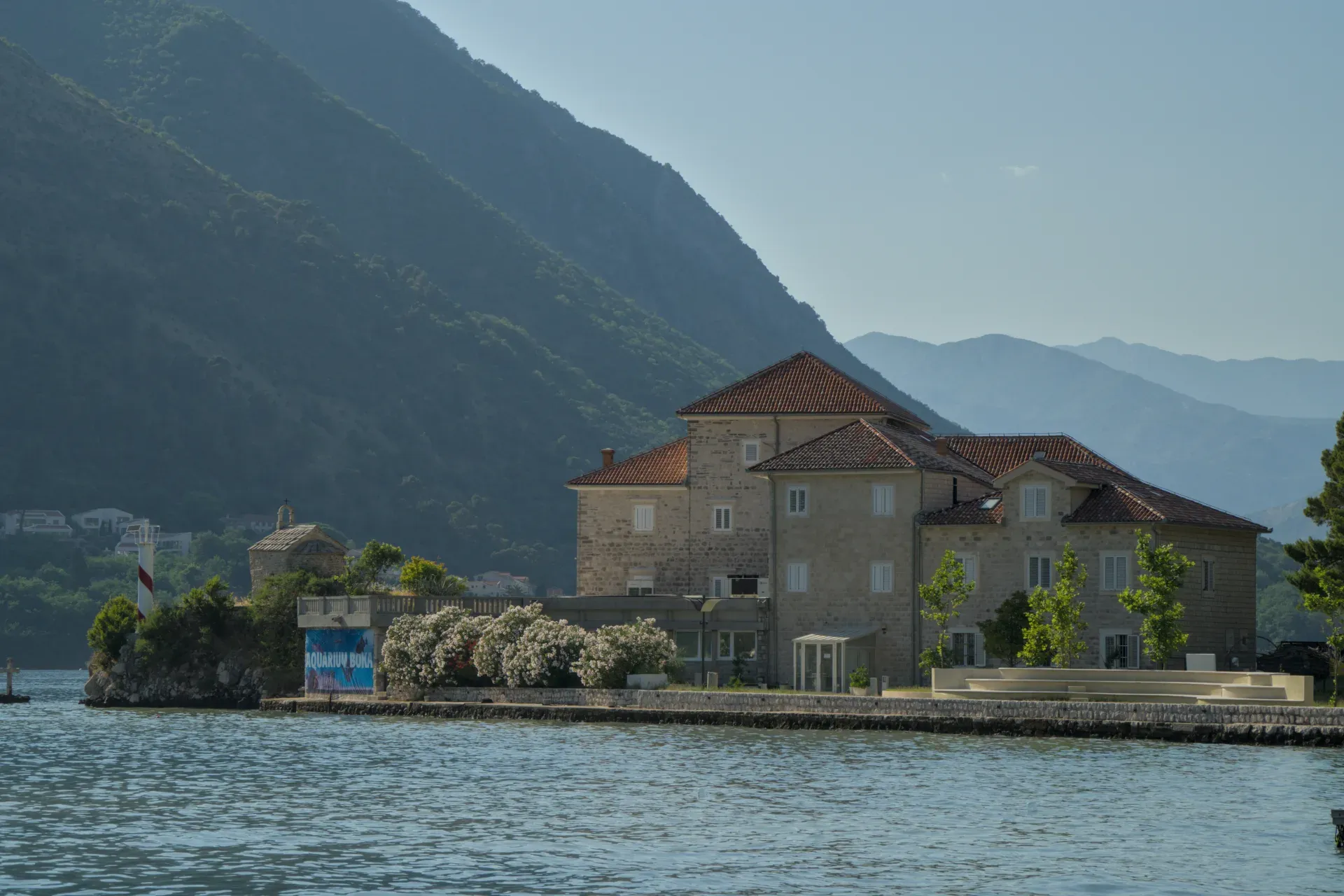 Edificio histórico en la Bahía de Kotor con montañas al fondo y una estructura similar a un faro.