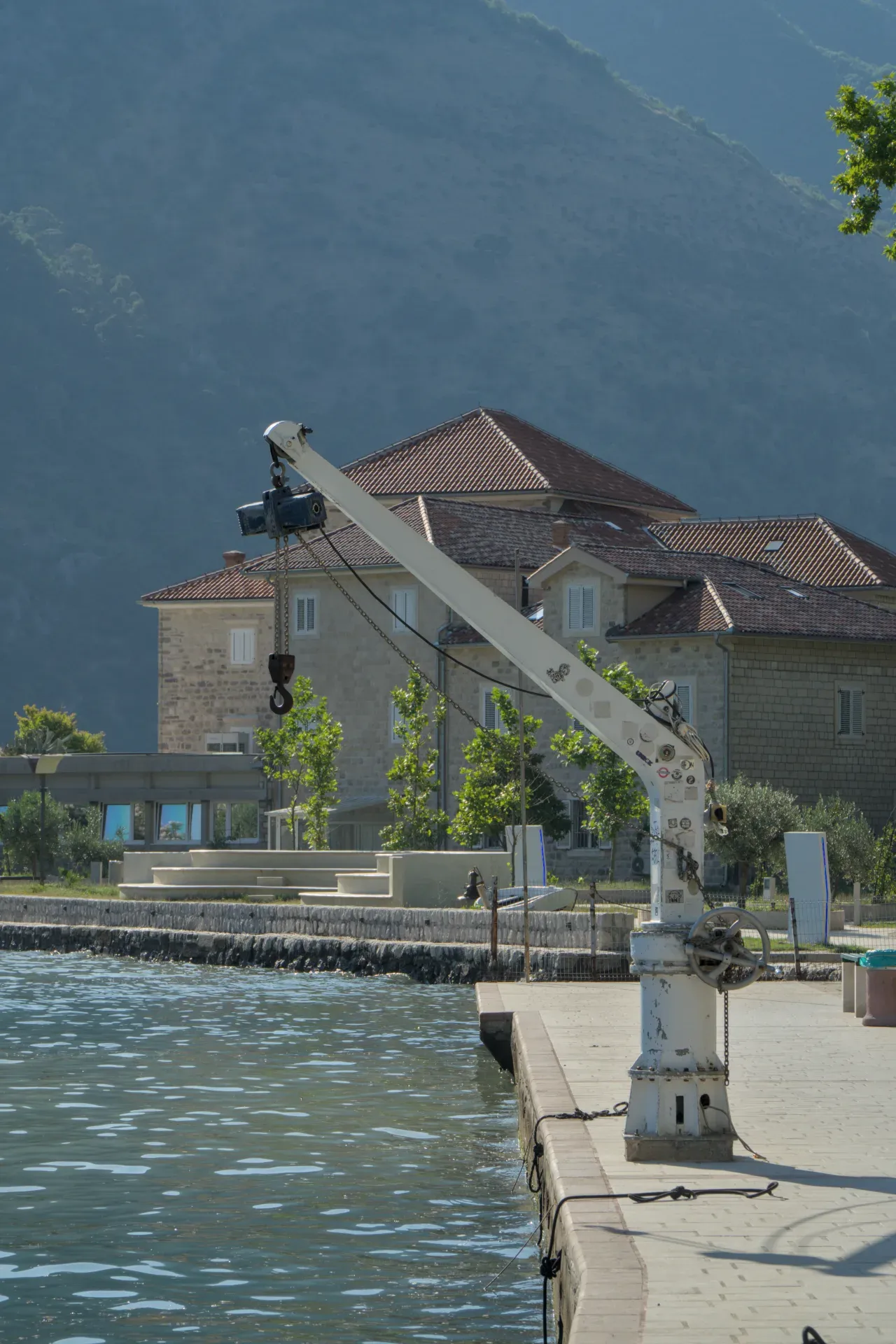 Une grue industrielle sur la rive de la baie de Kotor, avec une maison et une montagne en arrière-plan.