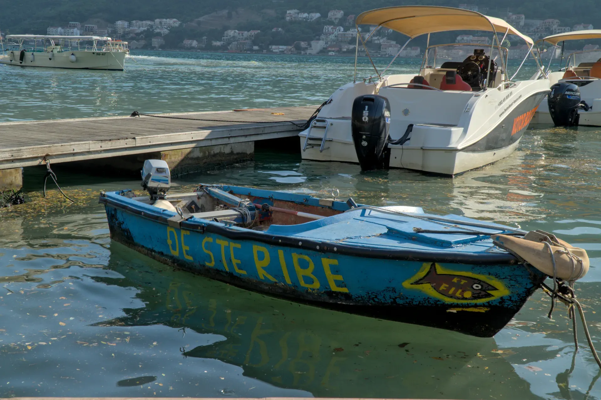 Altes blaues Fischerboot mit gelbem Schriftzug "DE STERIBE" und Fischsymbol, das in der Bucht von Kotor neben einem Steg und modernen Booten schwimmt.
