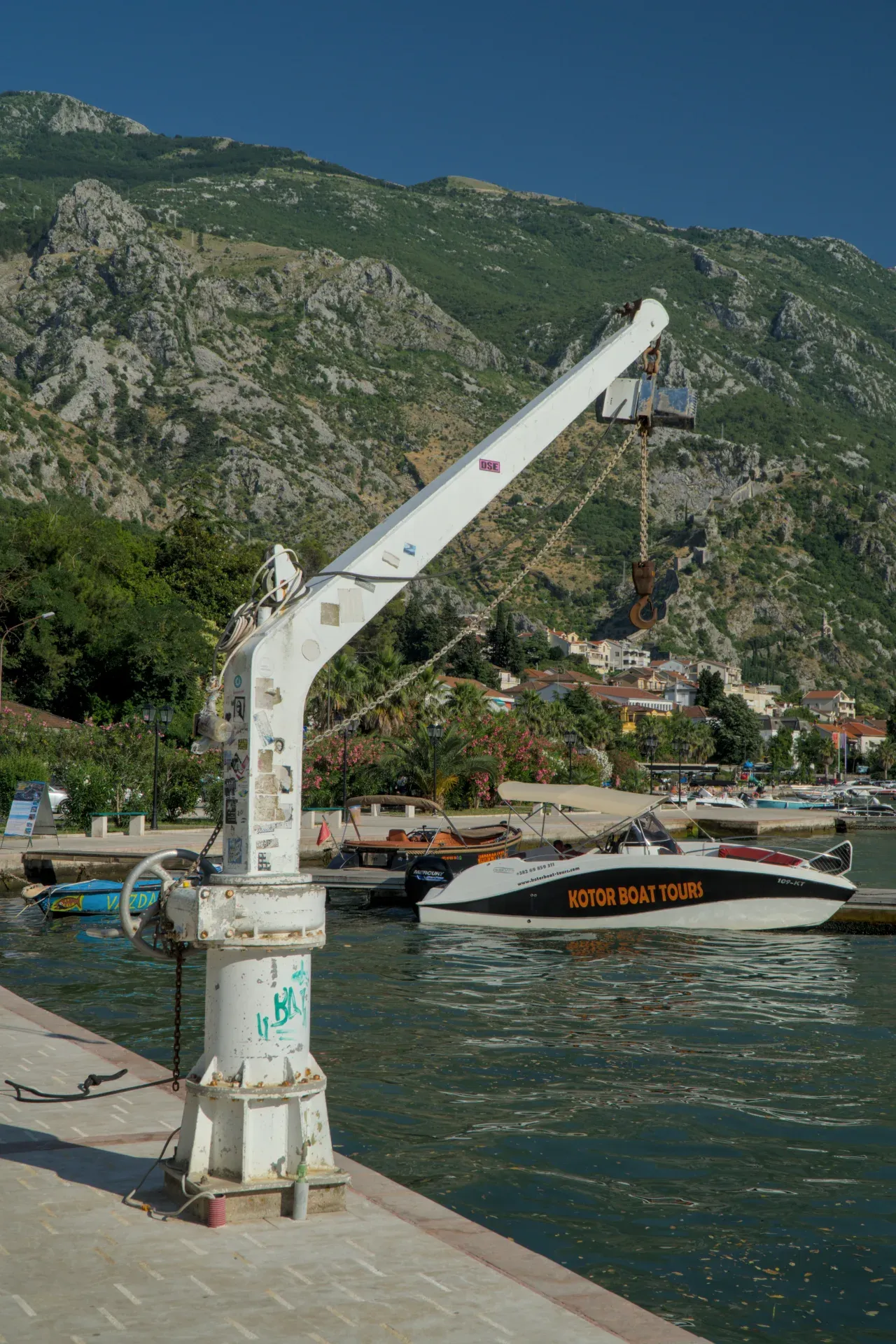 Une grue de port blanche se dresse au premier plan, avec des bateaux et la ville de Kotor à l'arrière-plan, devant un paysage montagneux.