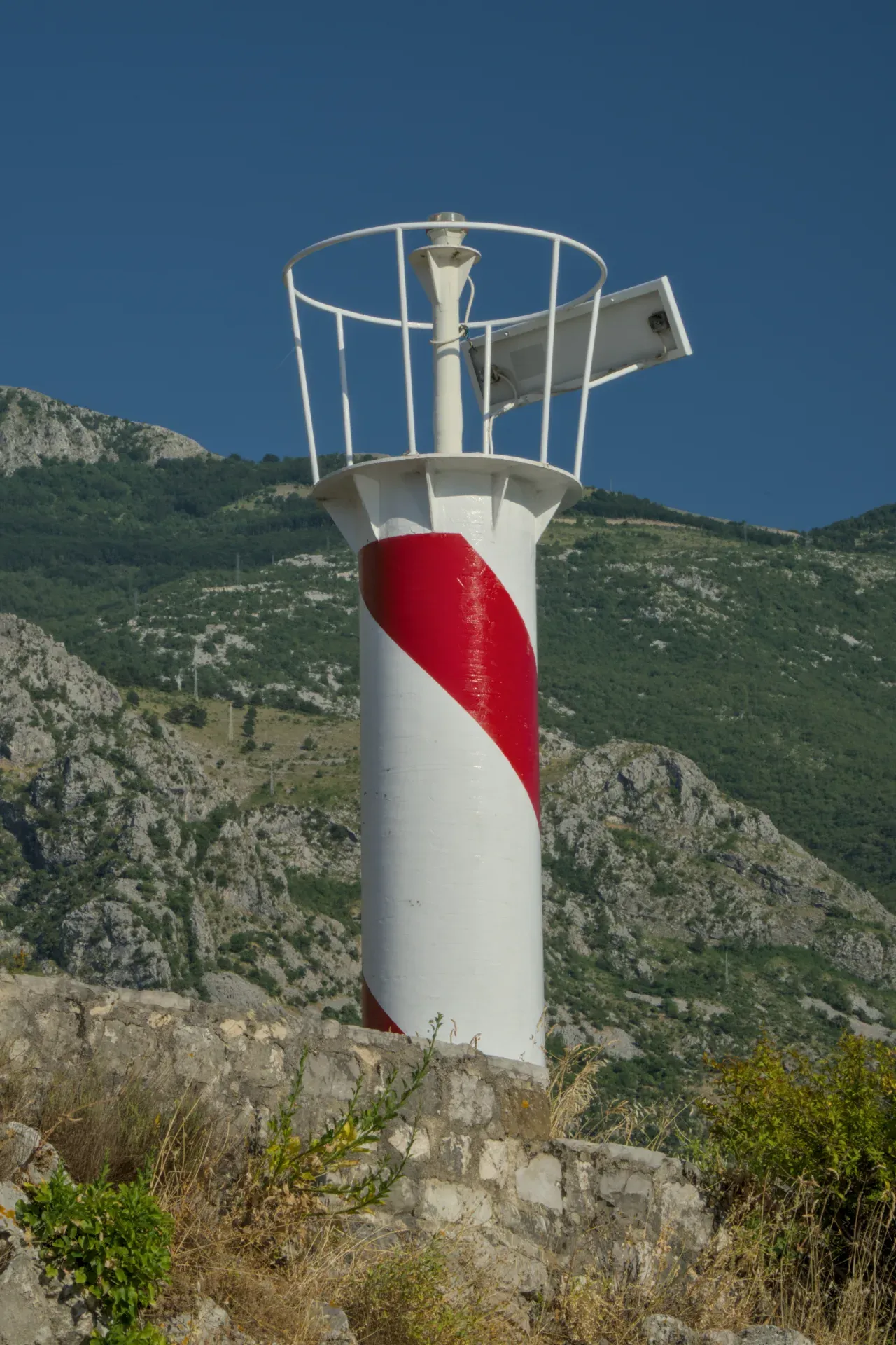 A striking red and white lighthouse stands on a rock against a blue sky and green mountains.