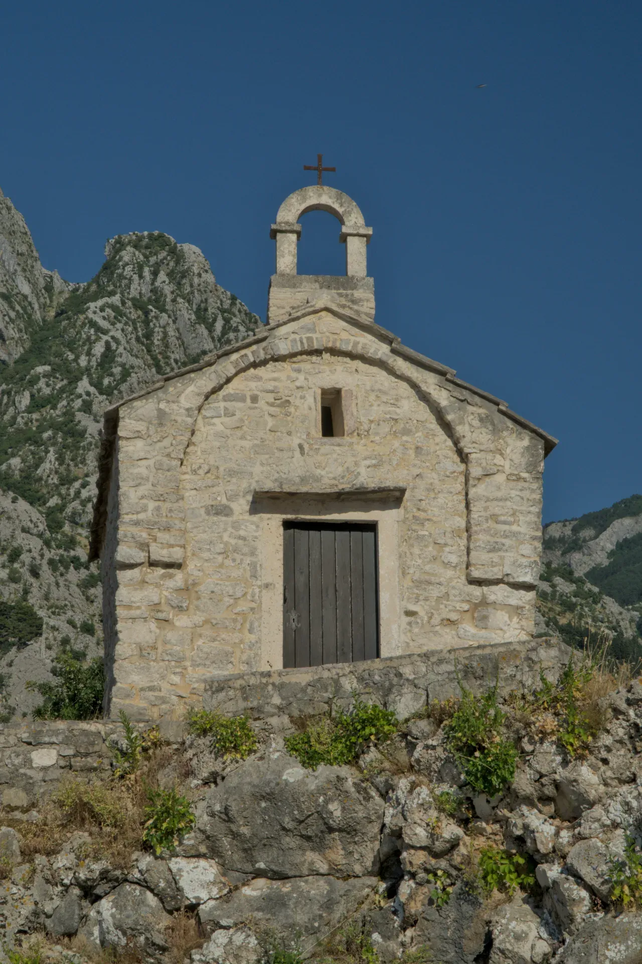 Die Kirche des Heiligen Elias, ein Steingebäude mit Holztür, steht auf einem felsigen Hang unter blauem Himmel in Dobrota, Montenegro.