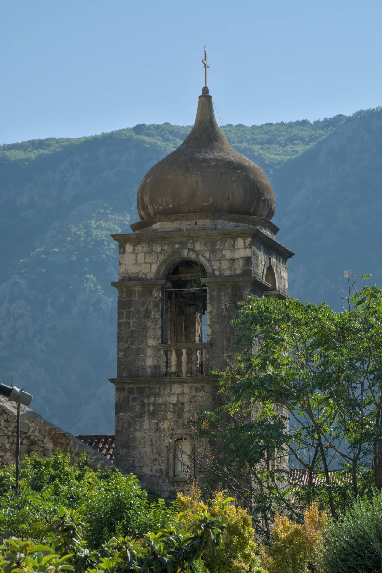Historic stone church tower with a dome and cross, set against forested mountains and a blue sky in Kotor.