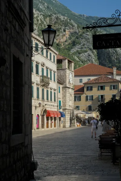 Malerei-artige Aufnahme einer Gasse in Kotor mit einem historischen Uhrturm, umrahmt von alten Gebäuden und Berglandschaft.