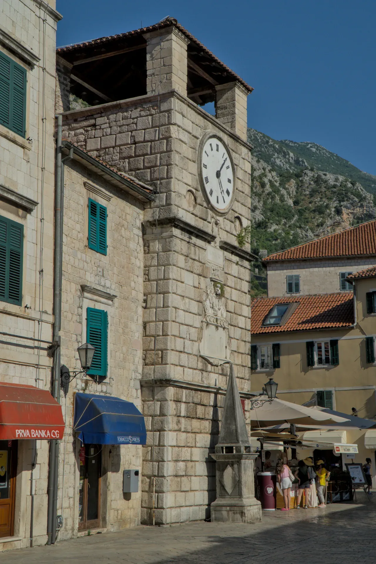 Ein steinerner Uhrturm in Kotor, Montenegro, mit grünen Fensterläden und einem Berg im Hintergrund.