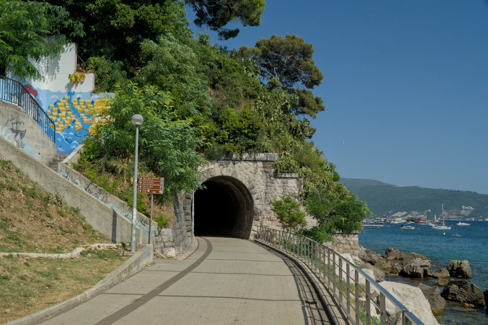 Un túnel de piedra con entrada iluminada conduce por la costa de Herceg Novi, Montenegro, con vistas al mar y a las montañas.