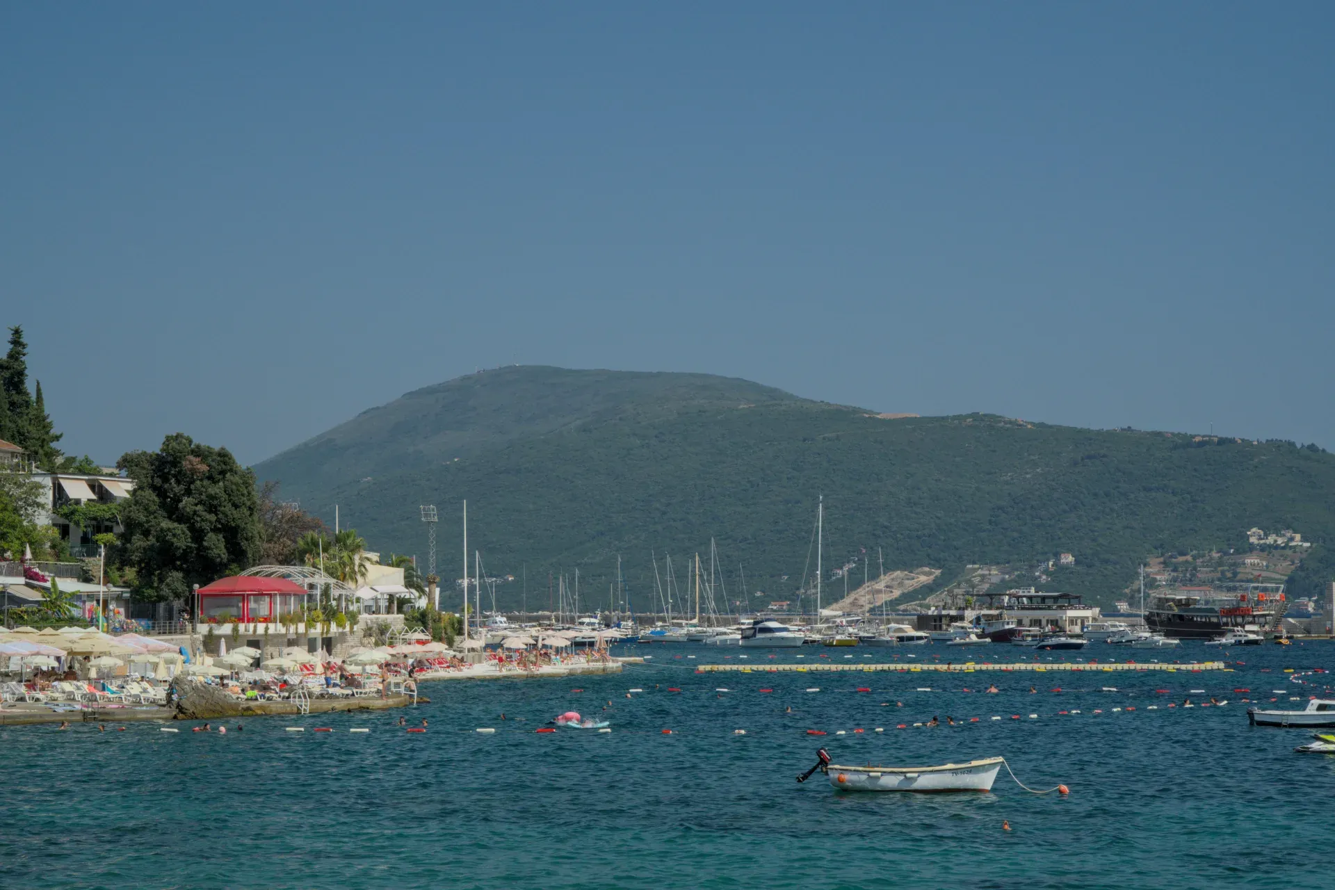 Brodovi u luci Herceg Novi, Crna Gora, sa živopisnim šetalištem i zelenim brdima u pozadini.