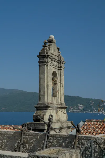 Un'antica torre dell'orologio in pietra si staglia contro un cielo blu intenso e il mare, con ancore storiche in primo piano.