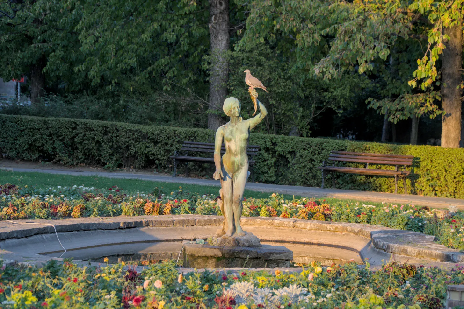 Statua di bronzo di una ragazza con una cornucopia e una colomba nel Parco del Danubio a Novi Sad, Serbia, con fiori e alberi sullo sfondo.