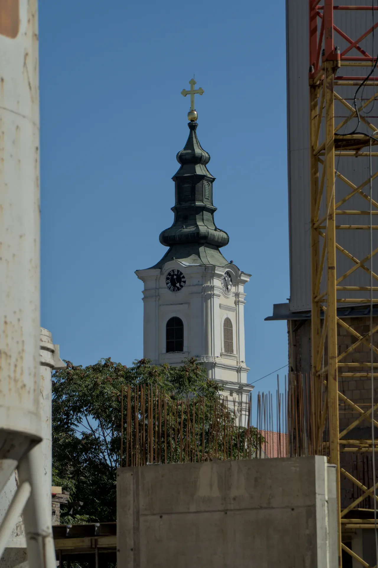 The bell tower of the Dormition Cathedral in Novi Sad rises against a clear blue sky, with its green roof and golden cross.