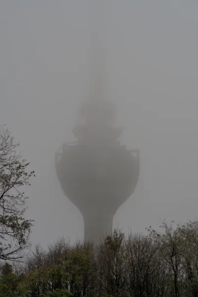 Una torre de televisión se eleva en una densa niebla en la montaña Fruška Gora, su silueta apenas visible, con árboles en primer plano.