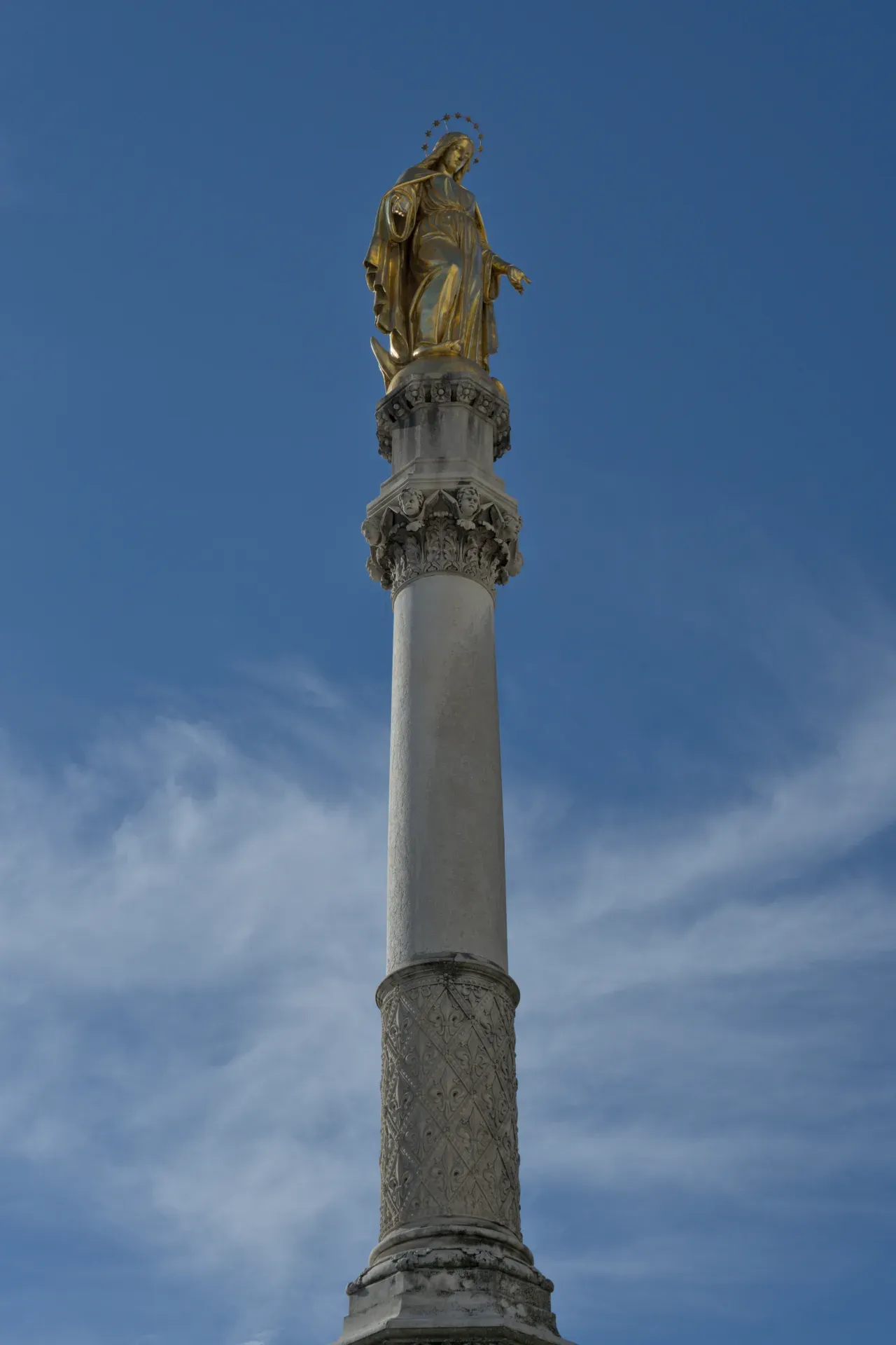 Goldene Marienstatue auf einer kunstvoll verzierten Säule vor einem blauen Himmel mit zarten Wolken.