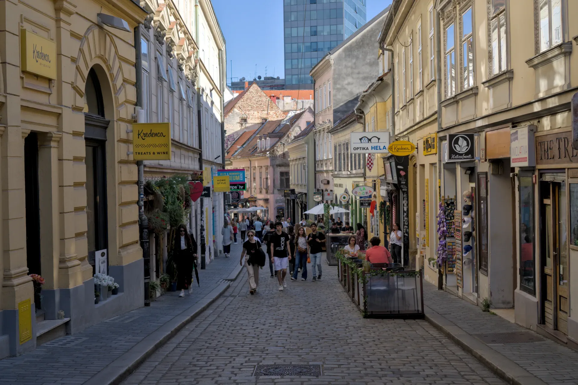Eine sonnenbeschienene, kopfsteingepflasterte Gasse in Zagreb, gesäumt von Geschäften und Cafés, mit Menschen, die spazieren gehen.