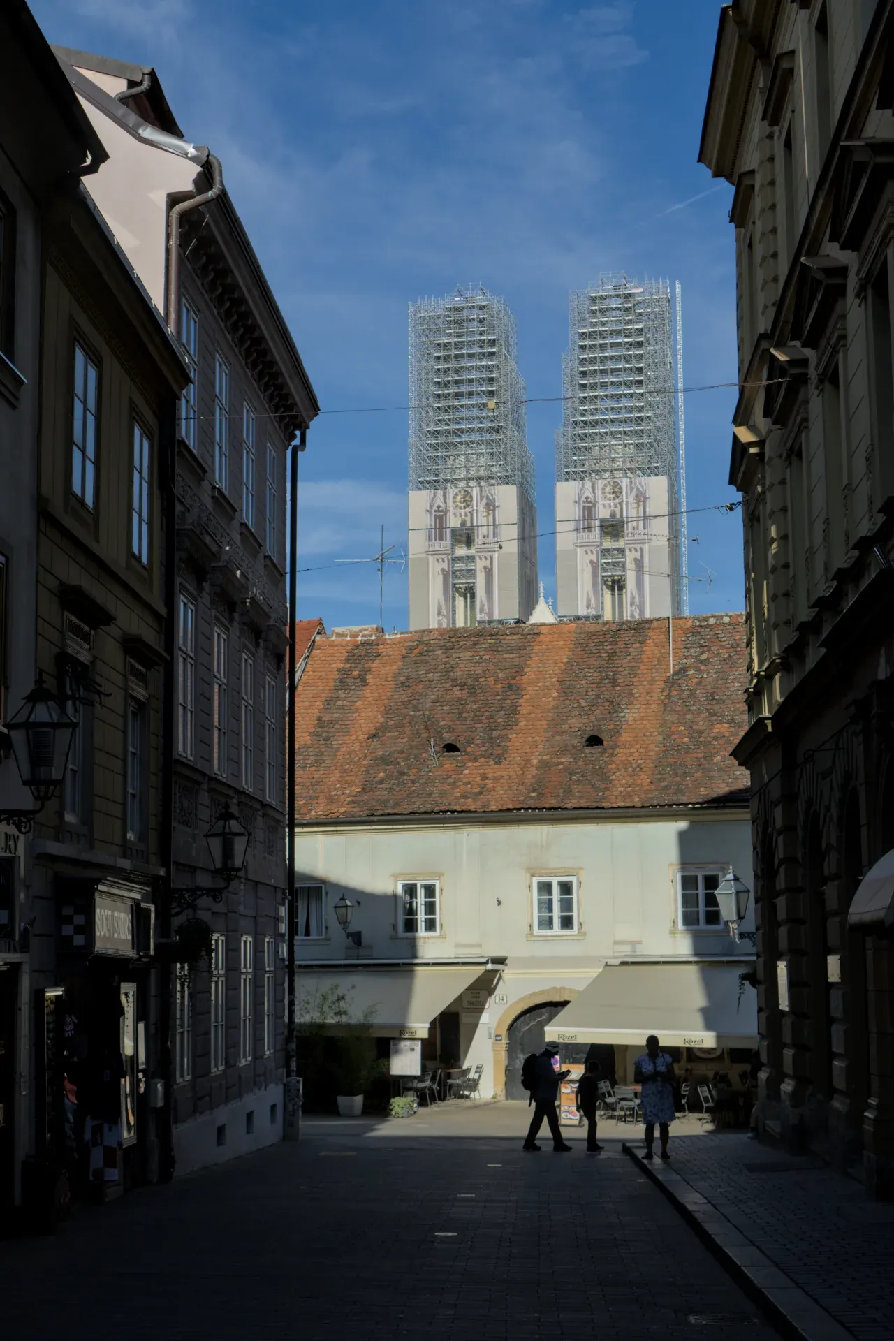 Architekturfotografie der Kathedrale von Zagreb, die im Gerüst vor blauem Himmel steht, mit Menschen, die durch eine Gasse gehen.
