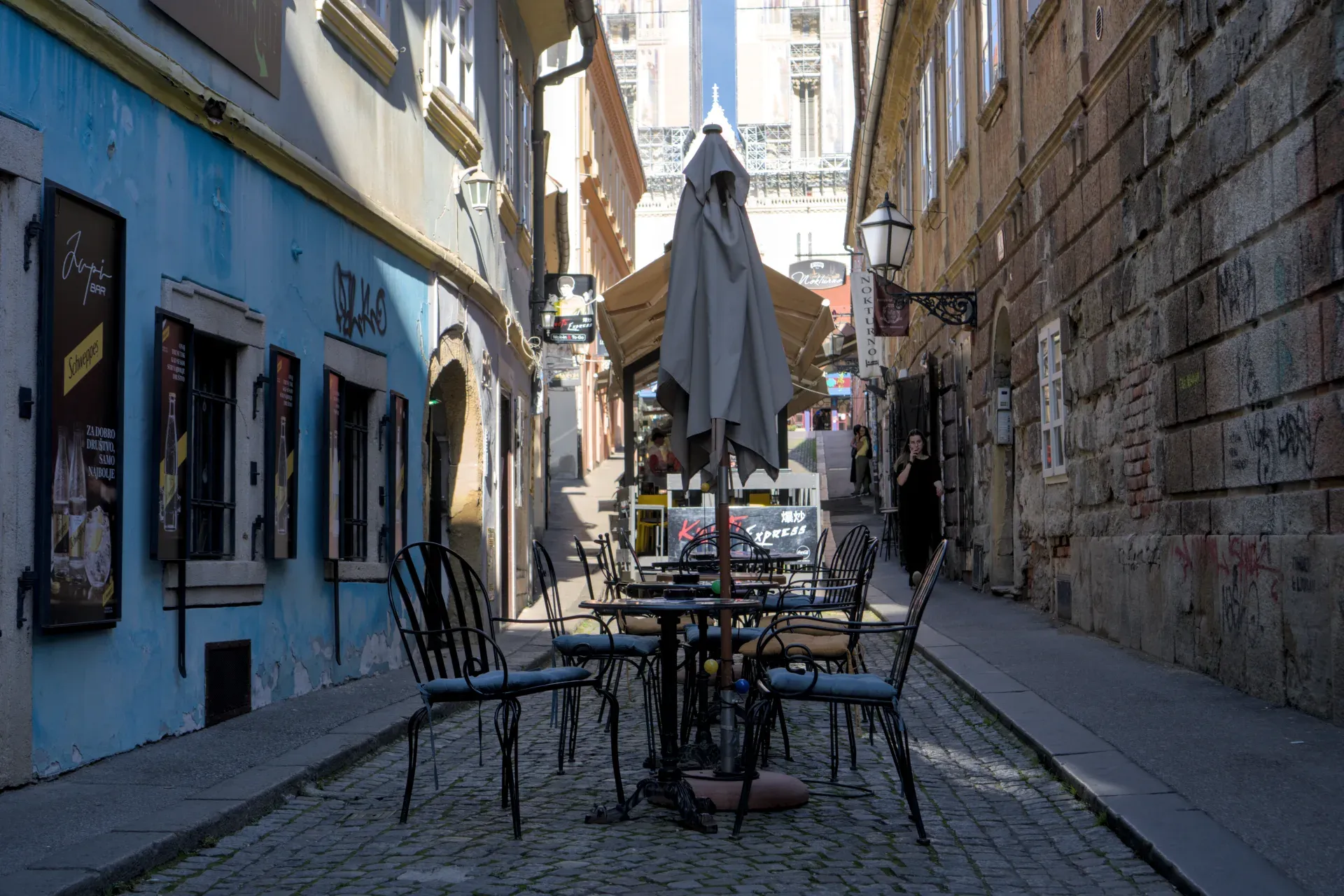 Uzak uličica u Zagrebu, obrubljena zgradama plave i kamene fasade, sa stolovima u kafiću u prvom planu.