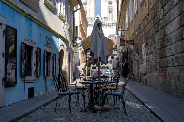 Eine schmale Gasse in Zagreb, gesäumt von Gebäuden mit blauer und steinerner Fassade, mit Cafétischen im Vordergrund.