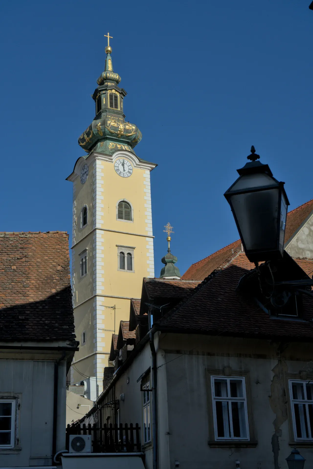 Portretna fotografija žute crkve sa zelenom kupolom i zlatnim ukrasima, snimljena na jasnom plavom nebu iznad Zagreba.