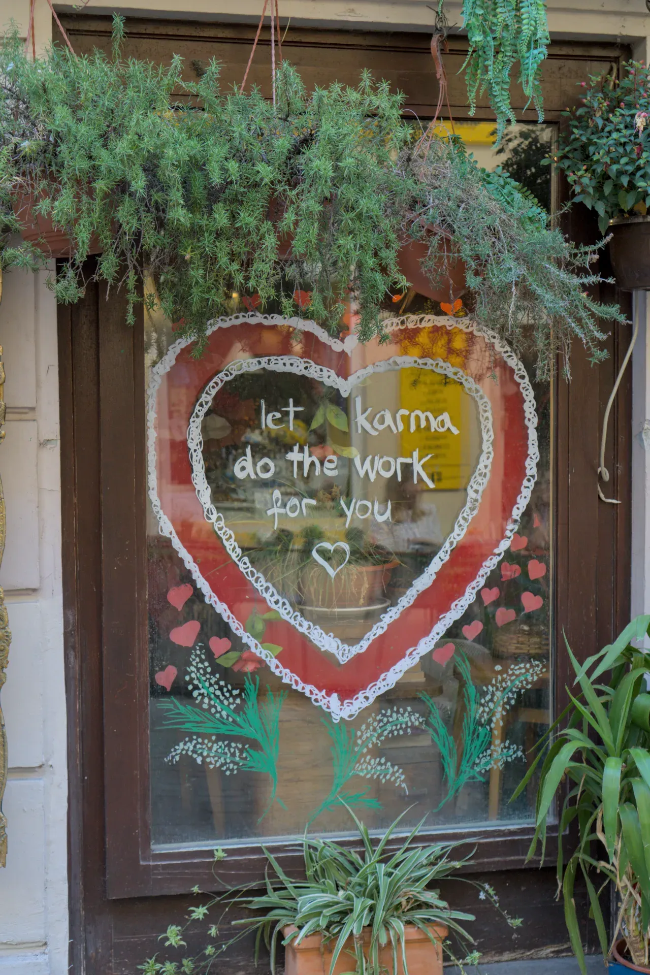 Un cuore dipinto a mano con la scritta 'Lascia che il karma faccia il lavoro per te' su una finestra, circondato da piante e accenti a forma di cuore.