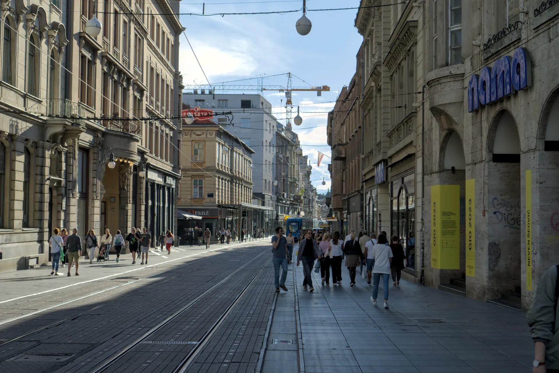 A sunny street in Zagreb with tram tracks and historic buildings, bustling with pedestrians.