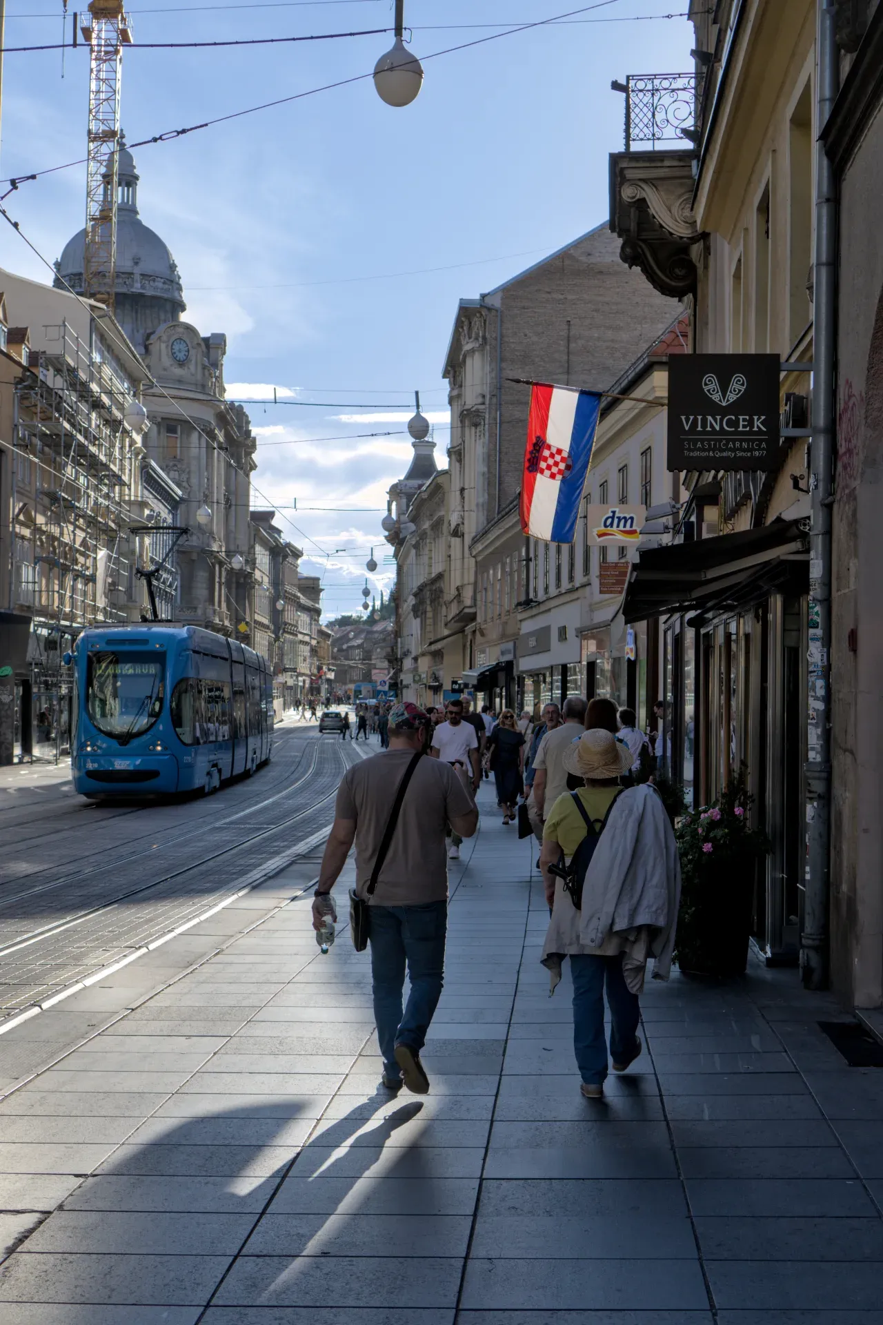 Un tramway bleu circule sur les voies dans une rue de Zagreb, bordée de bâtiments et de piétons.