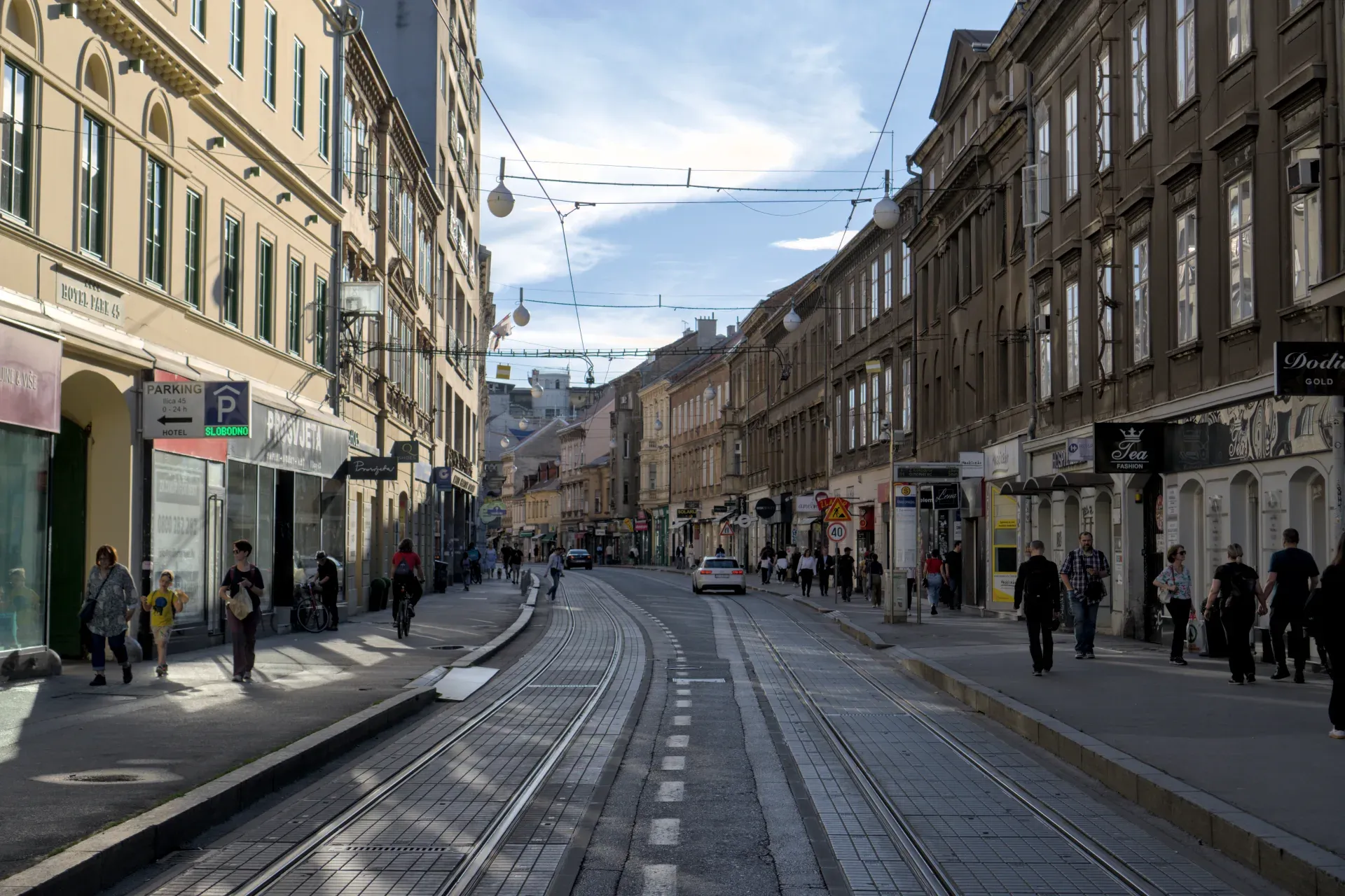 An urban street in Zagreb with historic buildings, tram tracks, and people walking on a sunny day.