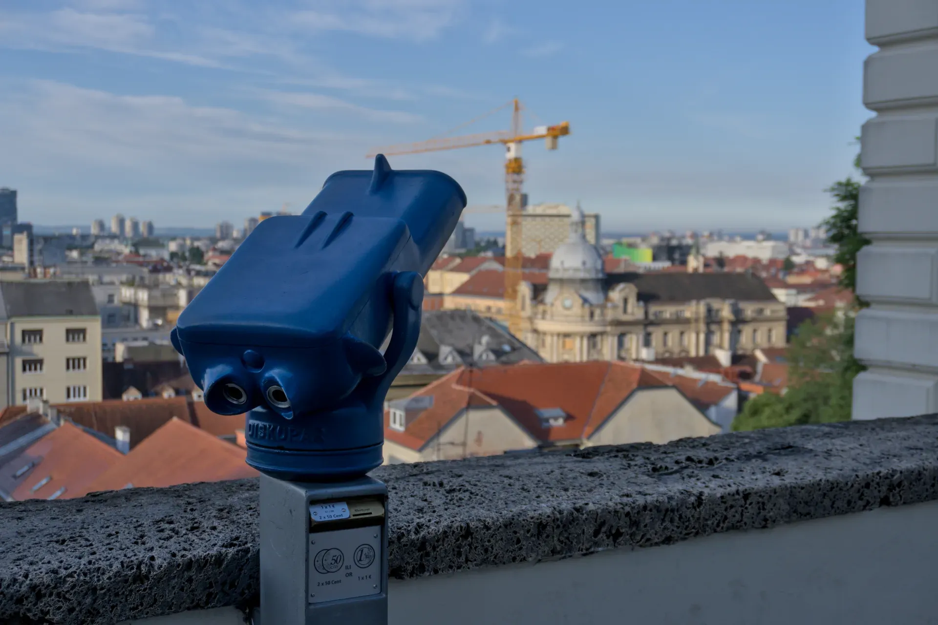 Un binocolo da osservazione blu si staglia davanti allo scenografico skyline di Zagabria, con tetti rossi e una gru sullo sfondo.