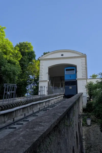 Die blaue Standseilbahn von Zagreb fährt bergauf auf ihren Schienen vor einem weißen Gebäude und blauem Himmel.