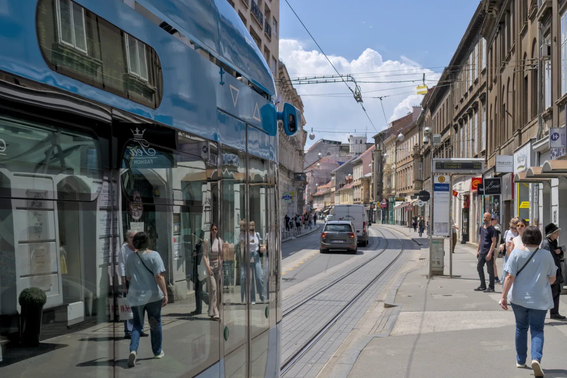Un tram blu si muove per la strada trafficata di Zagabria, riflettendo edifici e persone, con architettura storica sullo sfondo.