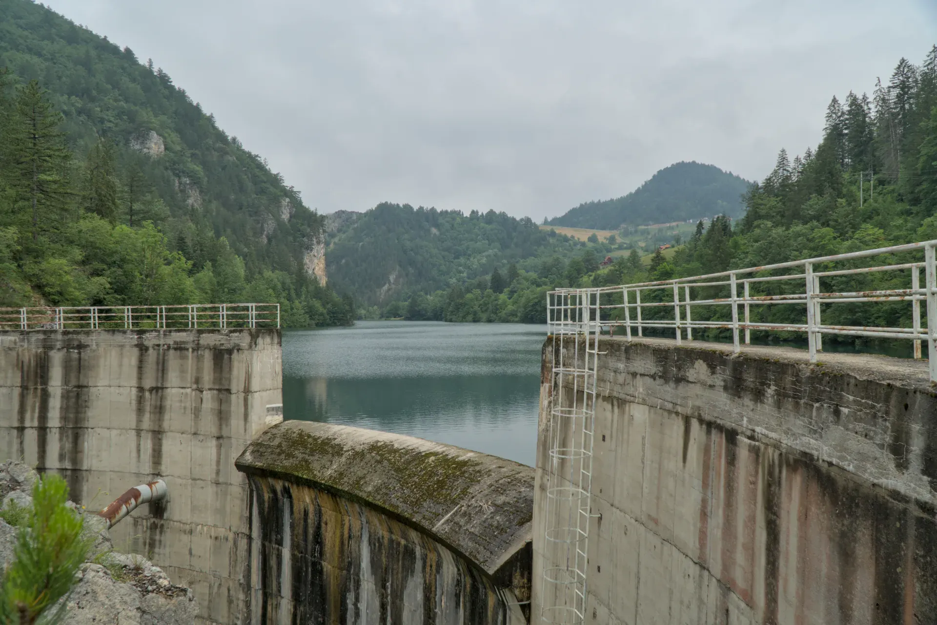 Der Damm am Spajića See in Serbien, umgeben von bewaldeten Bergen, mit ruhigem Wasser.