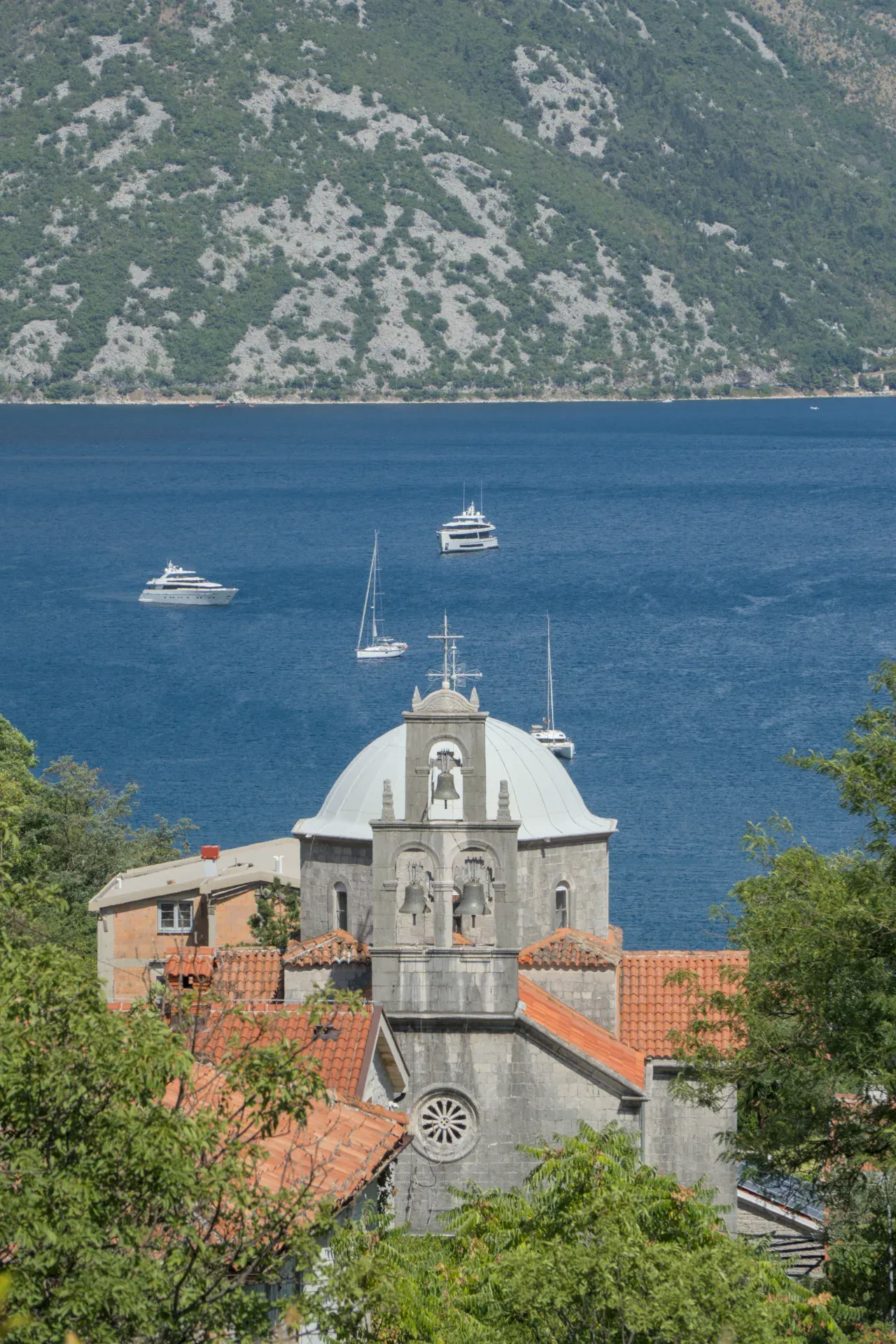 Panoramablick auf die Bucht von Kotor in Morinj, Montenegro, mit einer Kirche und Segelbooten auf dem blauen Wasser.