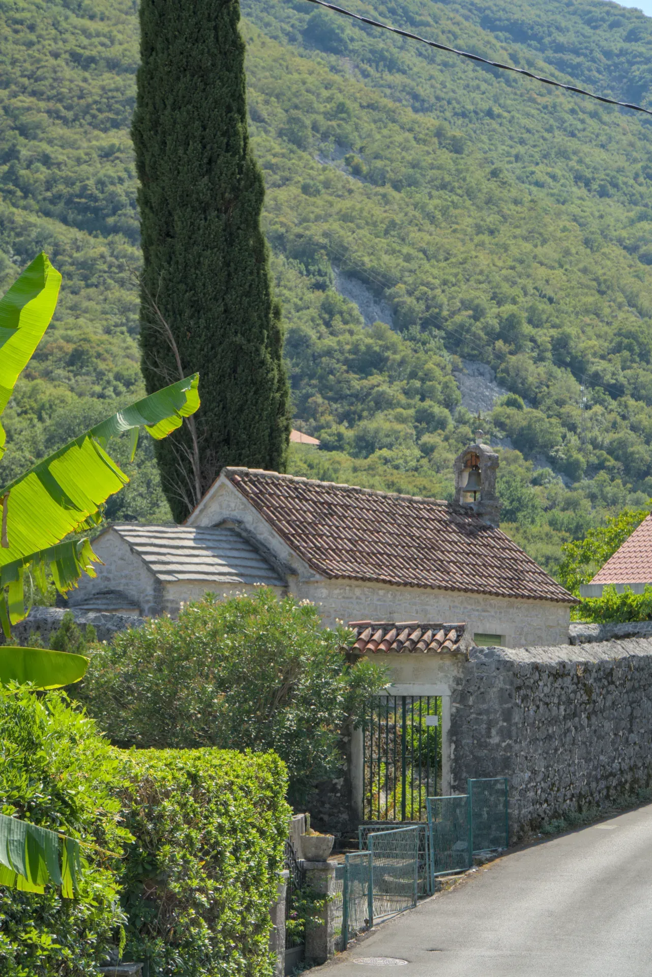 Una piccola chiesa in pietra con tetti di tegole in terracotta, fiancheggiata da vegetazione lussureggiante e un alto cipresso, vicino a Kotor, Montenegro.