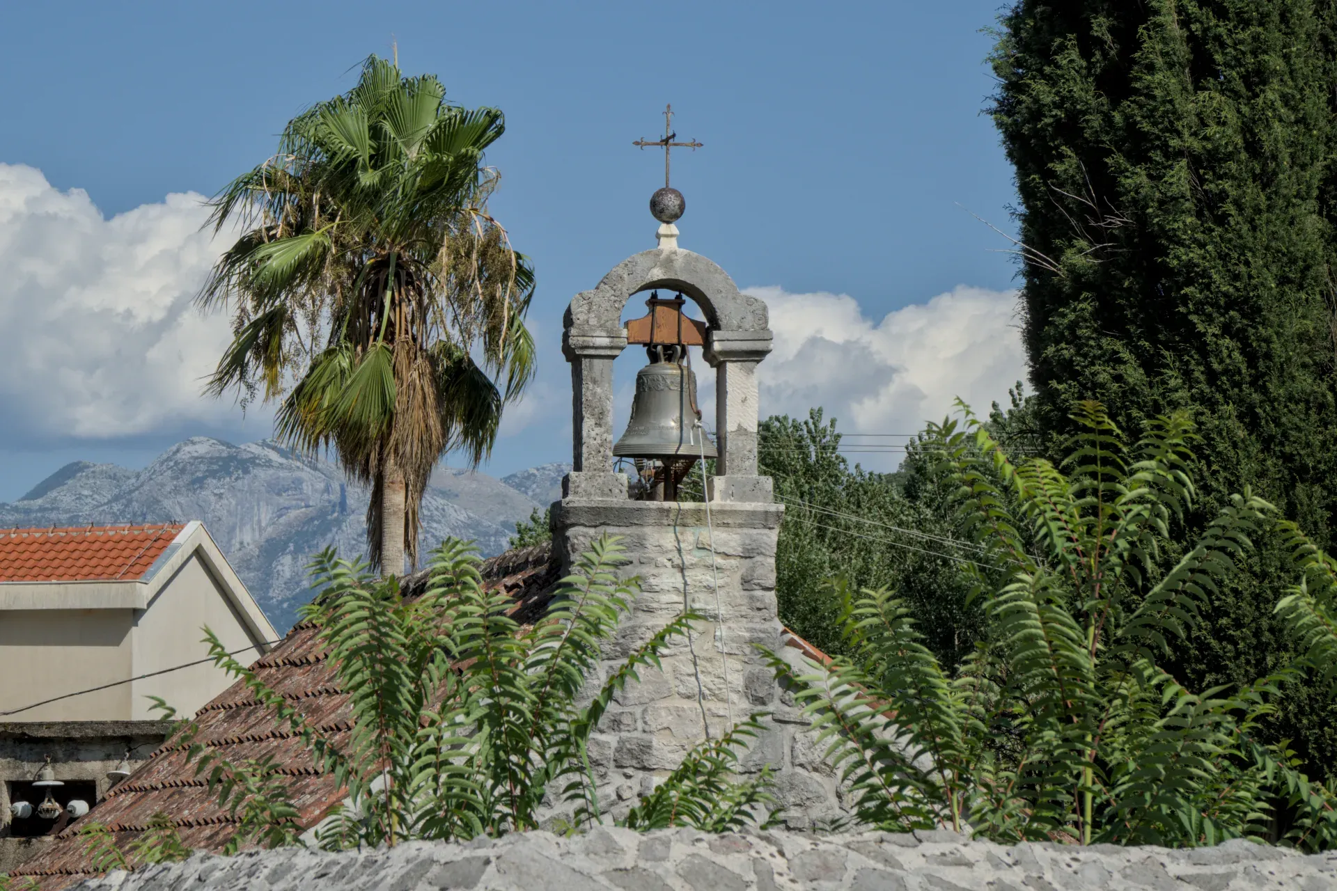 The "колокольня церкви Святого Тривуна" bell tower in Kotor, Montenegro, stands under a bright blue sky with mountains behind.