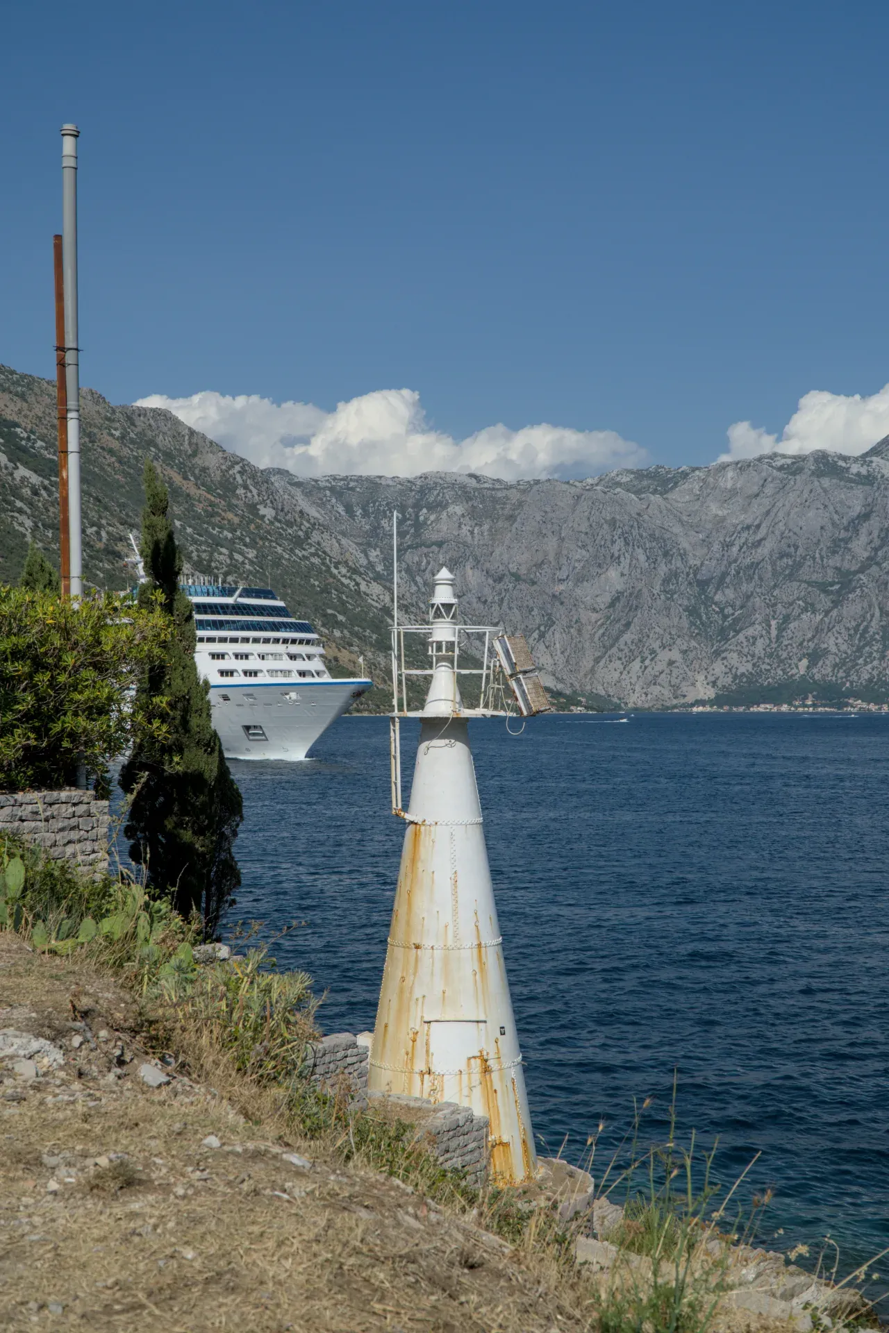 Un faro bianco stagionato si erge sulla riva della Baia di Kotor, con una grande nave da crociera che naviga sull'acqua.
