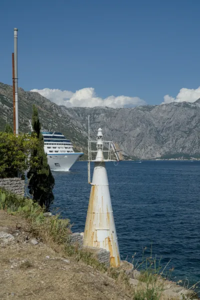 Un faro bianco stagionato si erge sulla riva della Baia di Kotor, con una grande nave da crociera che naviga sull'acqua.