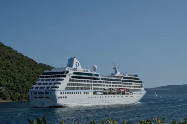 Ein großes weißes Kreuzfahrtschiff verlässt die Bucht von Kotor unter blauem Himmel, die Hügel Montenegros im Hintergrund.