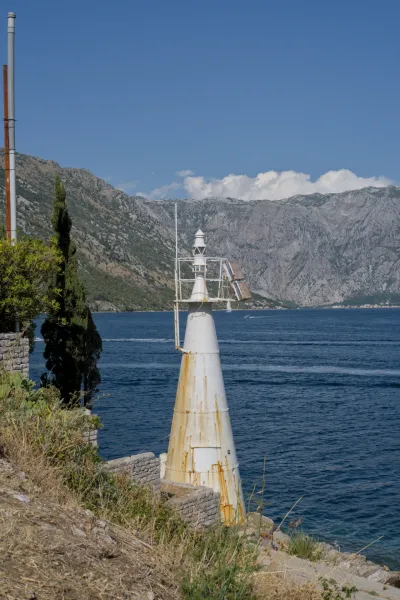 Faro bianco stagionato all'ingresso della Baia di Cattaro, Montenegro, con montagne e mare blu sullo sfondo.