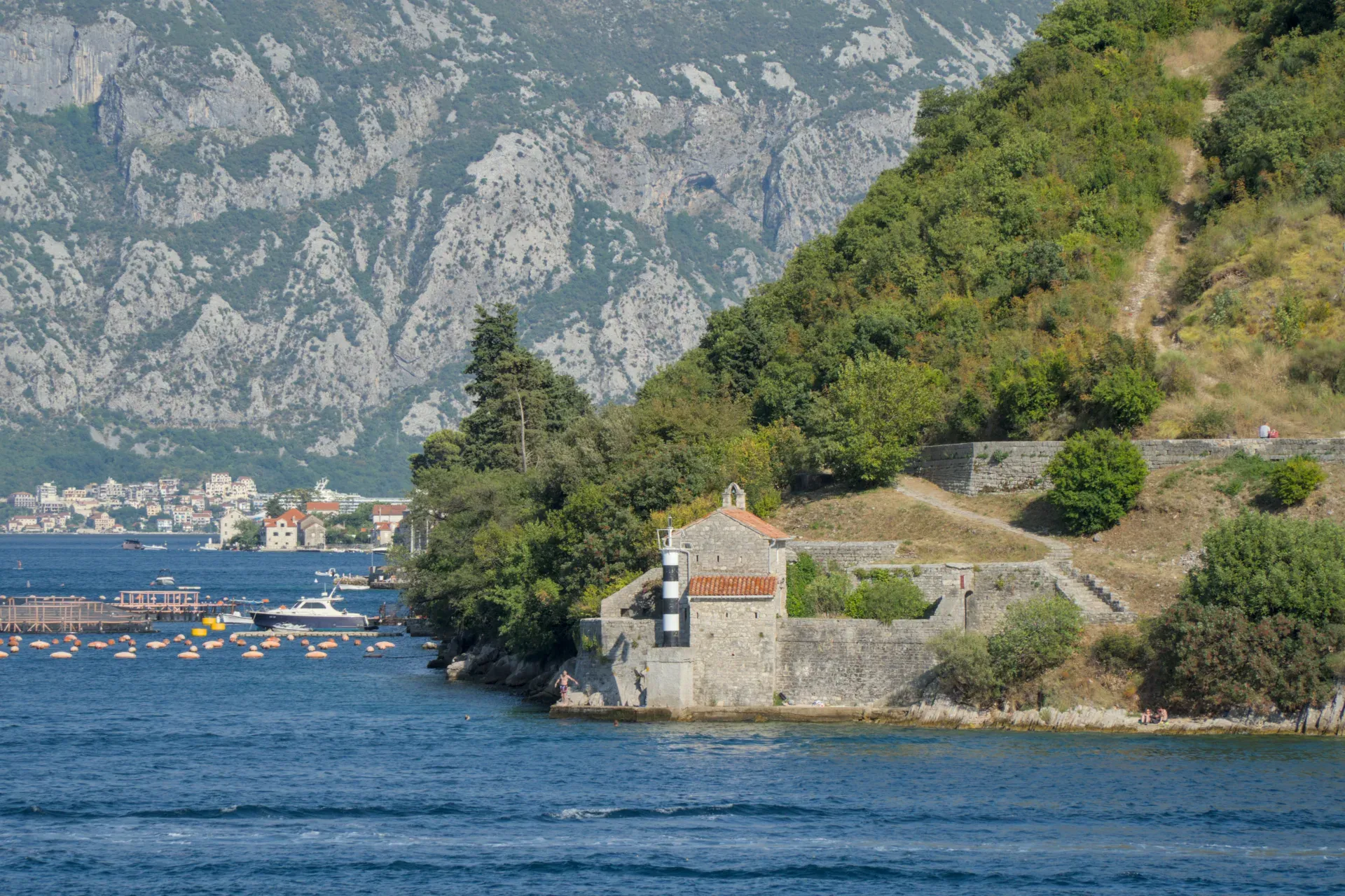 Una piccola cappella in pietra con un faro a strisce bianche e nere sulla costa, di fronte a montagne e acqua blu a Kotor.