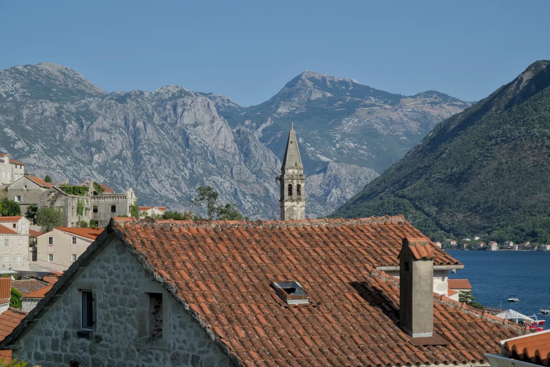 Perast, Montenegro: Ein Blick auf die städtischen Dächer vor einer Bergkulisse mit einem Kirchturm und dem Wasser.