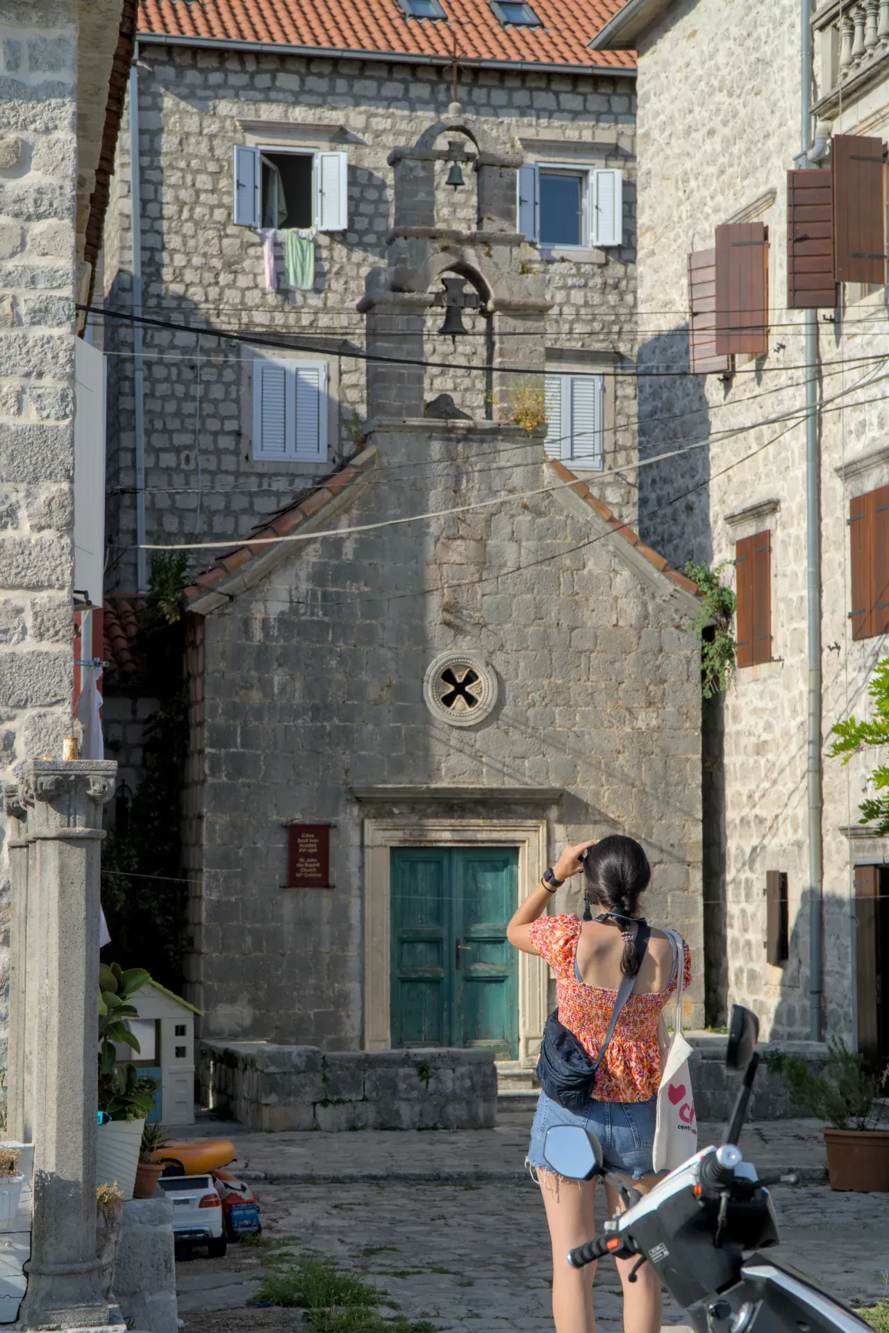 Une femme photographie la "Ruelle de Perast" à Kotor, Monténégro, avec une façade d'église en pierre et une architecture vénitienne.