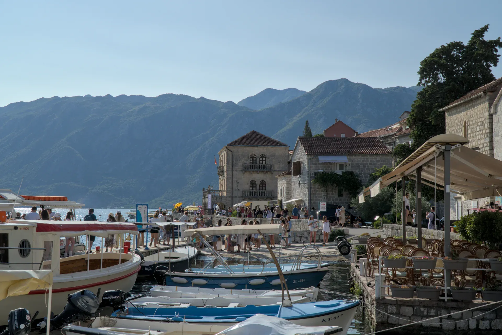 Ein sonniger Tag am Kai von Perast, Montenegro, mit Booten, historischen Gebäuden und Bergen im Hintergrund.