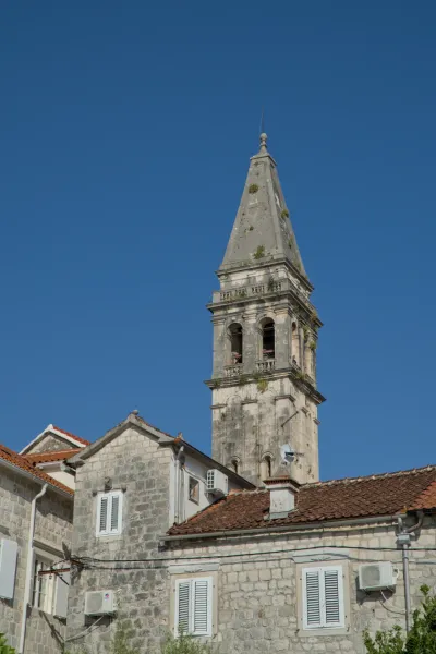 El campanario de piedra de Perast se alza contra un cielo azul claro, flanqueado por casas tradicionales.
