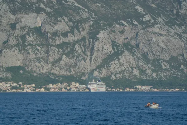 Ein Kreuzfahrtschiff und kleine Boote auf dem Meer vor der Stadt Kotor mit ihren Häusern und den dahinter liegenden Bergen.