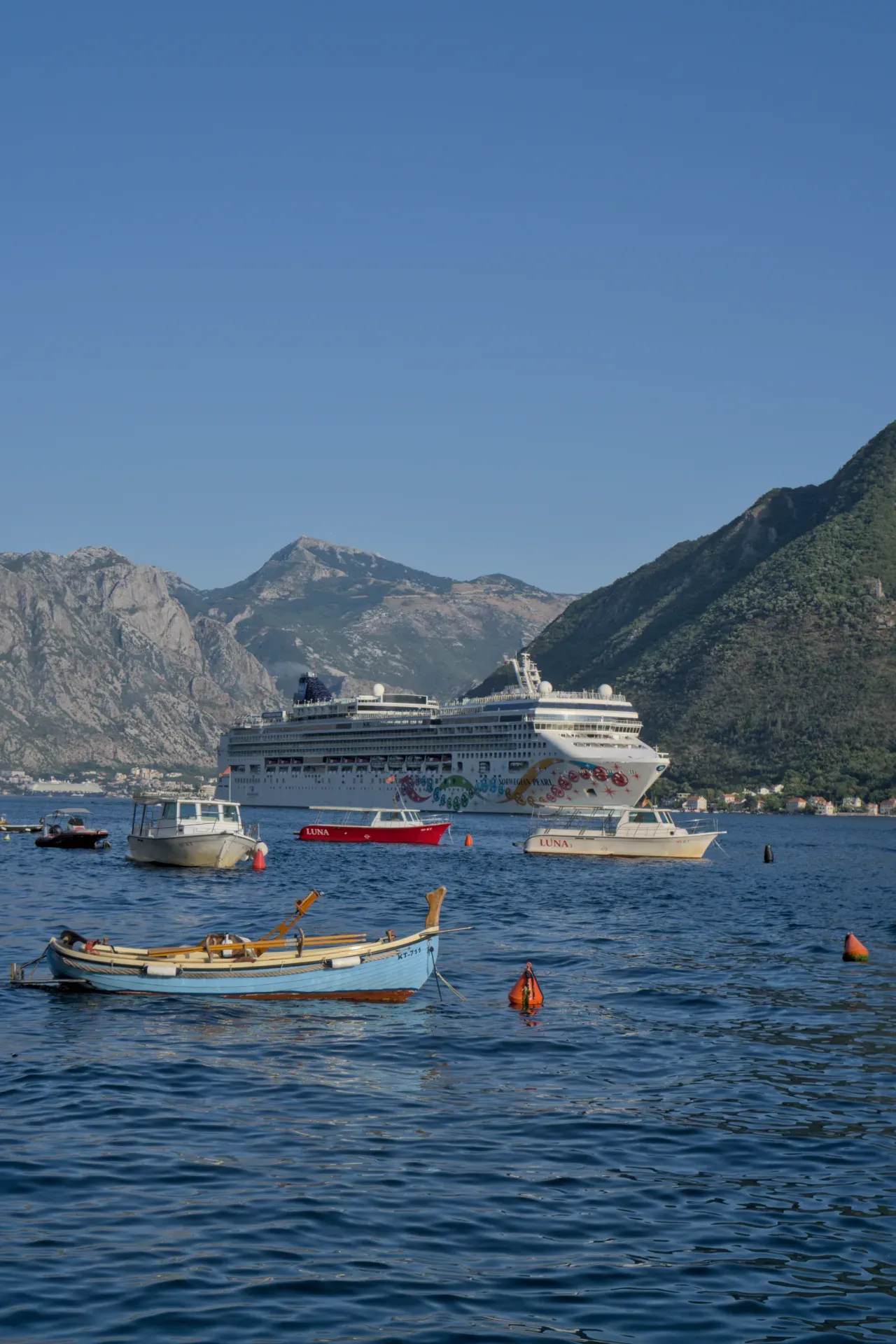 Veliki kruzer napušta Bokokotorski zaliv kroz Perast, sa manjim čamcima u prvom planu.