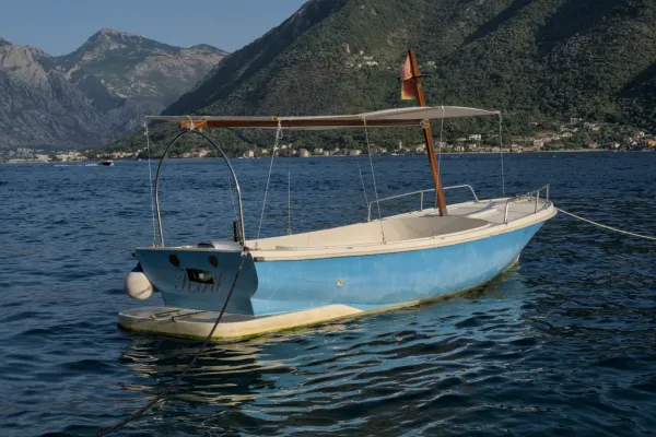 Un barco azul está amarrado en el agua frente a la costa de Perast, Montenegro, con montañas y casas al fondo.