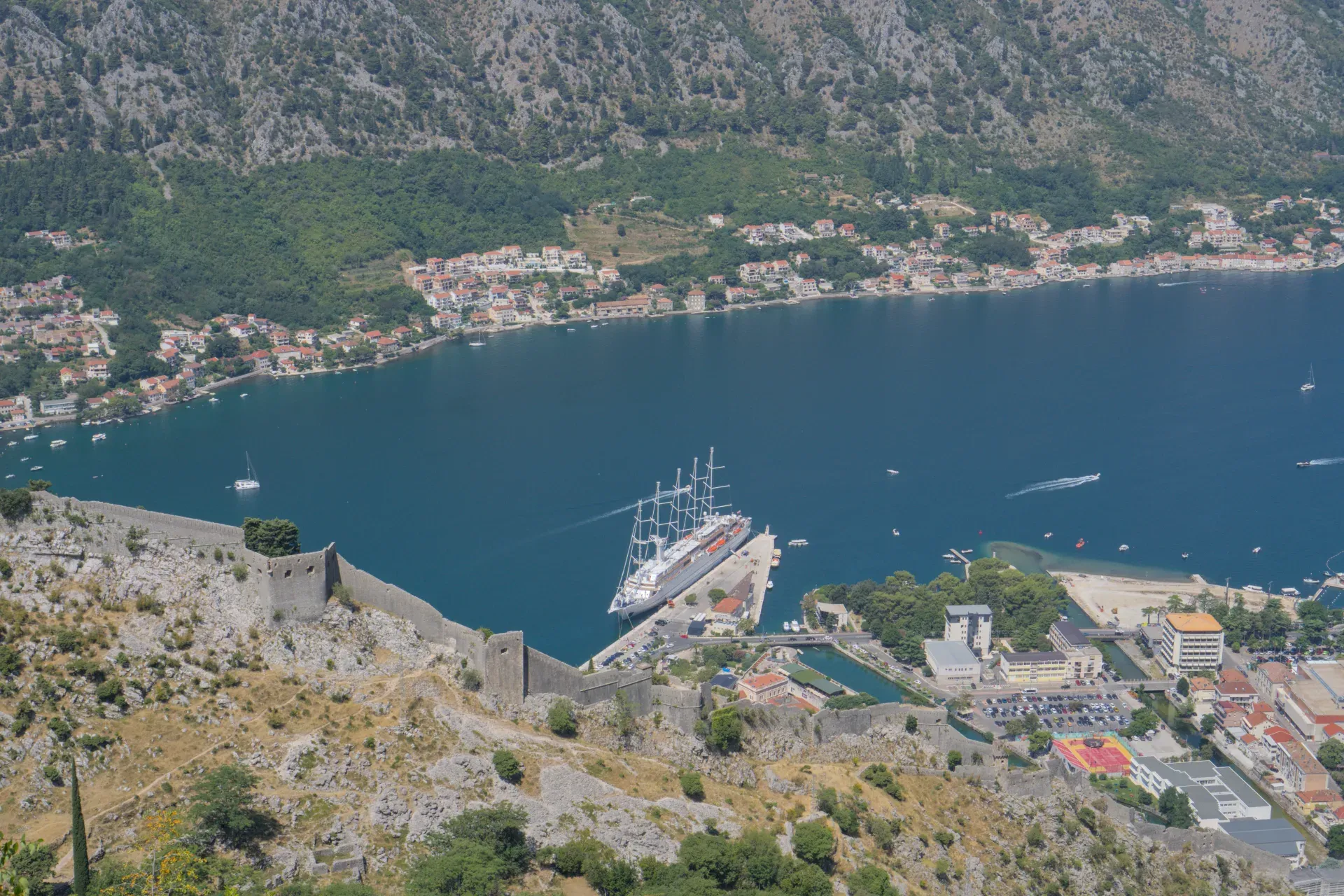 Un grande veliero è ormeggiato al molo nella baia di Kotor, Montenegro, con antiche fortificazioni in primo piano.